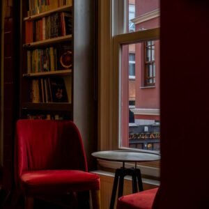 A cozy indoor space featuring red chairs, bookshelves, and a window view of the street outside.